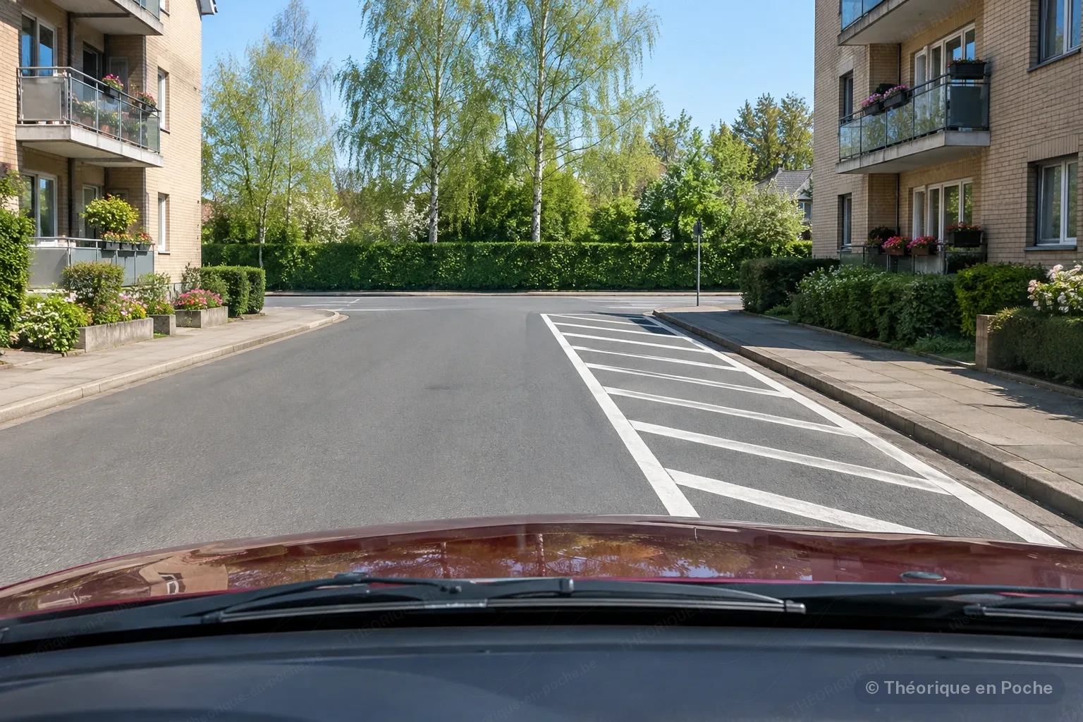 Image originale Tout pour mon permis (générée par IA, panneaux officiels) Driver's view approaching a T-junction between two brick buildings: on the right of the carriageway, an area of white diagonal stripes bordered by a solid line; on the left, the lane is clear.